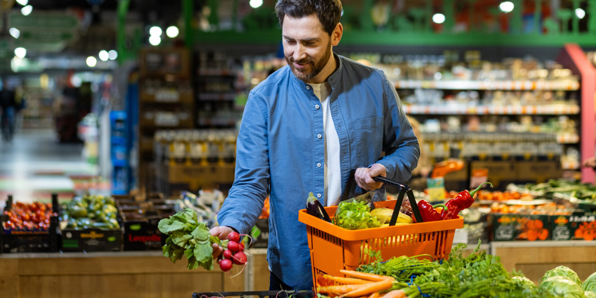 Man selecting fresh vegetables in grocery store, holding orange basket, surrounded by vibrant produce. Concept of healthy lifestyle and eco-friendly shopping in local market. Focus fresh food choice