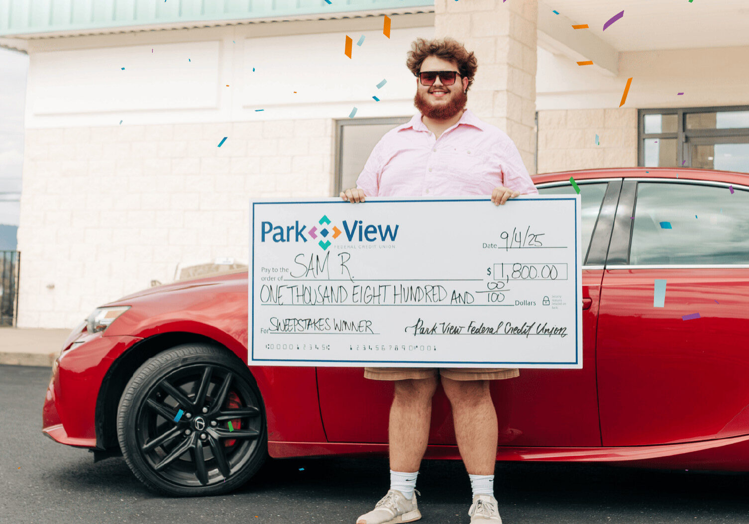 A man in sunglasses and casual clothes stands in front of a red car, holding a large check for $1,800 from Park View with confetti falling around him.