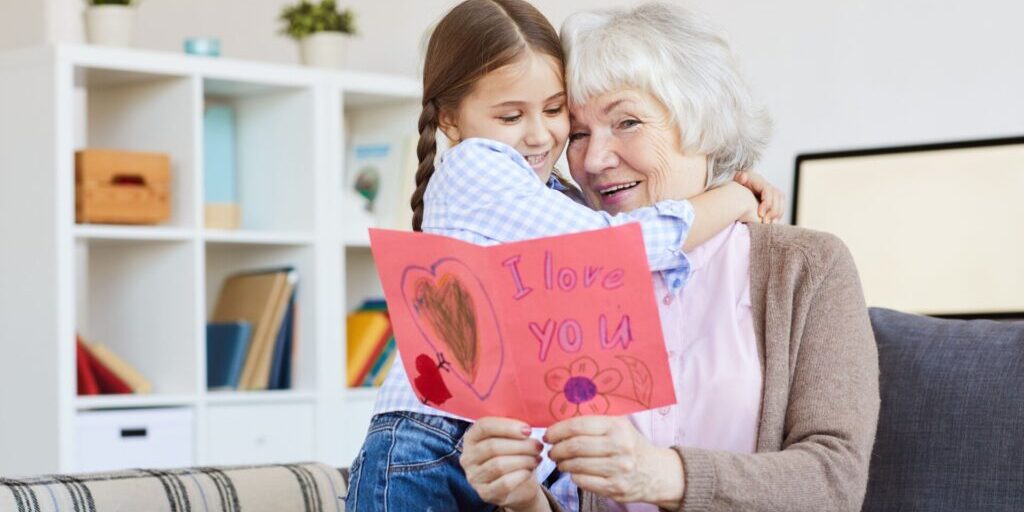 Granddaughter hugs her grandmother while giving a handmade Valentine’s card, a budget-friendly Valentine’s Day idea.
