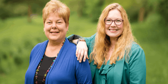 Two female Financial Advisors at Park View Federal Credit Union stand together smiling in a lush green park. One has short hair and a blue top, the other has long blonde hair and glasses, exuding warmth and friendship.