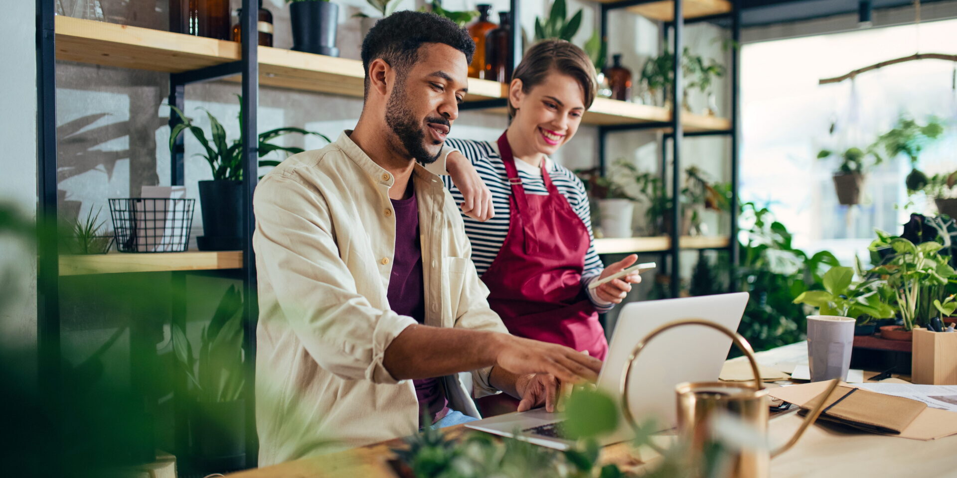 Smiling café coworkers review a tablet beside potted plants and a watering can in a bright, modern greenhouse-style workspace.