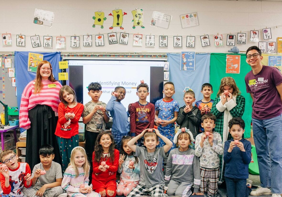 Cheerful classroom group portrait of a teacher and diverse children posing beneath alphabet decorations, with a learning screen behind them.