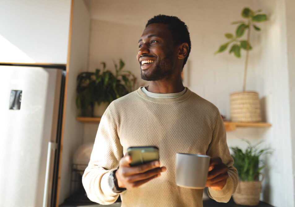 Happy African American Man Using Smartphone And Drinking Coffee In Kitchen Happy African American Man Using Smartphone And Drinking Coffee In Kitchen