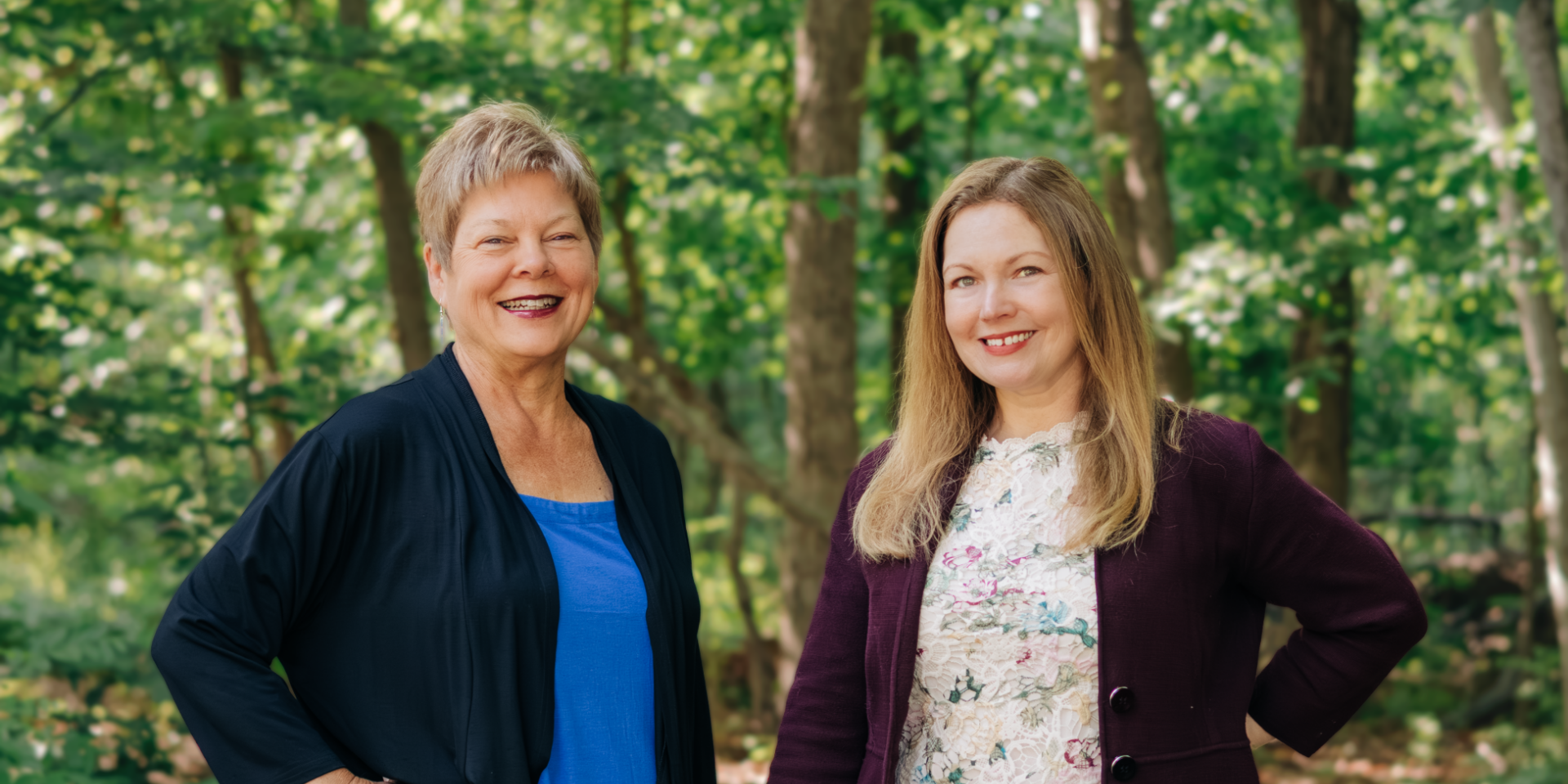 Two women smiling outdoors in a lush green forest, showcasing friendship and positivity in a natural setting.