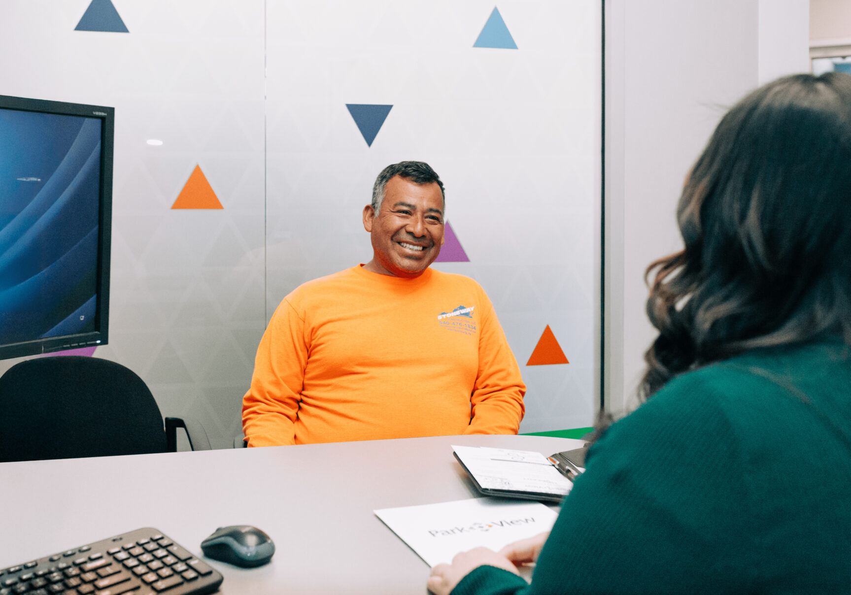 Smiling, middle-aged man in orange shirt sits at desk across from a Park View employee opening a Park View checking account.