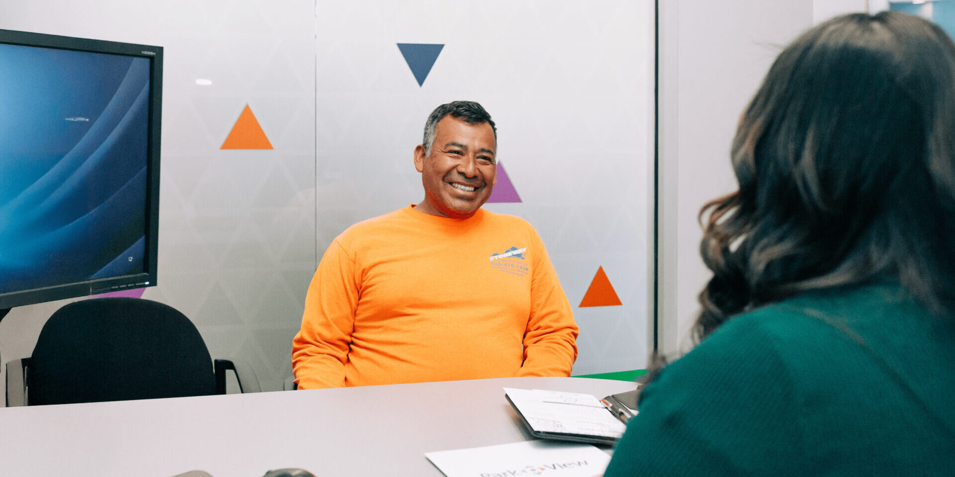 Smiling, middle-aged man in orange shirt sits at desk across from a Park View employee opening a Park View checking account.