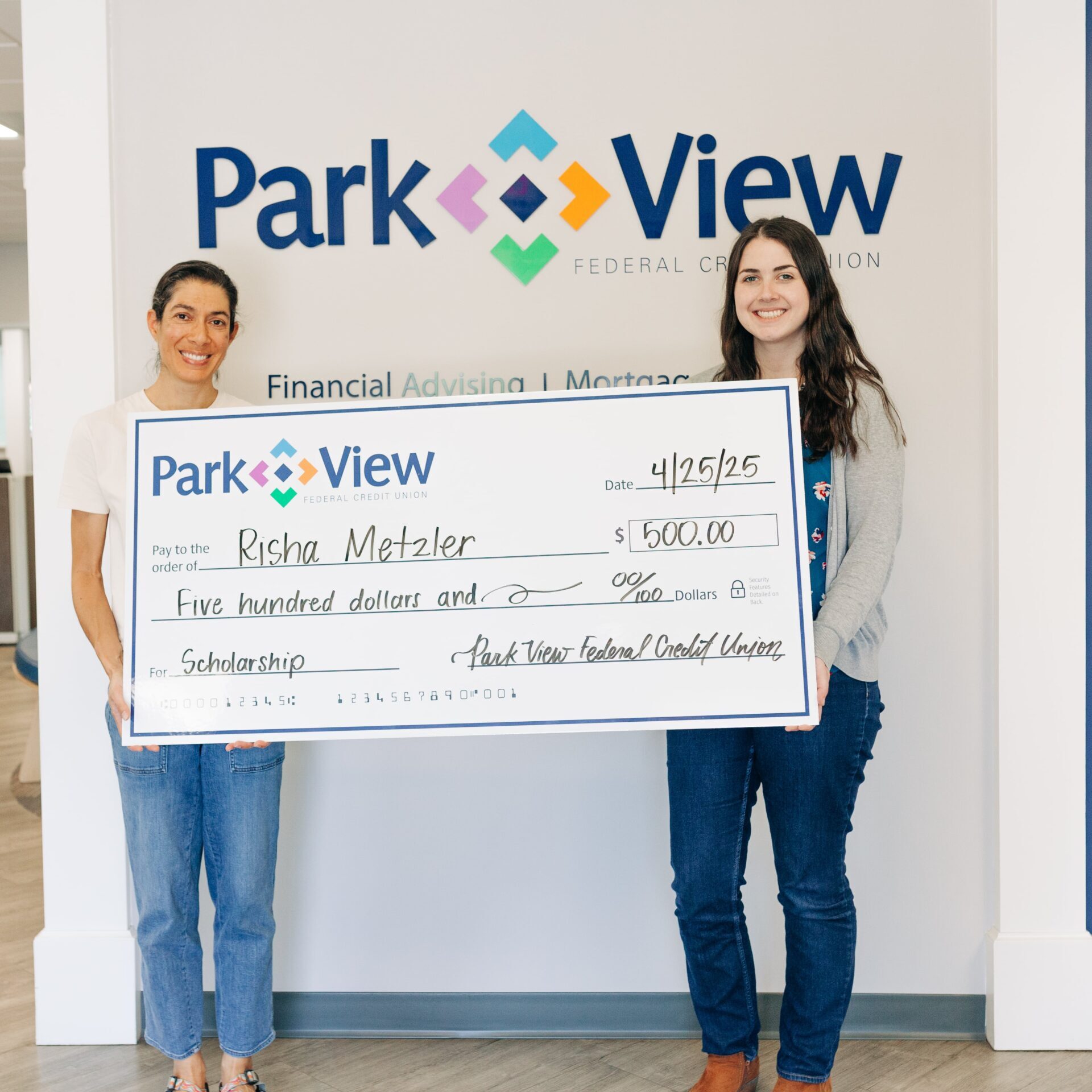 Two smiling people hold a $500 Park View Federal Credit Union scholarship check, posing before a branded wall in a cheerful presentation.