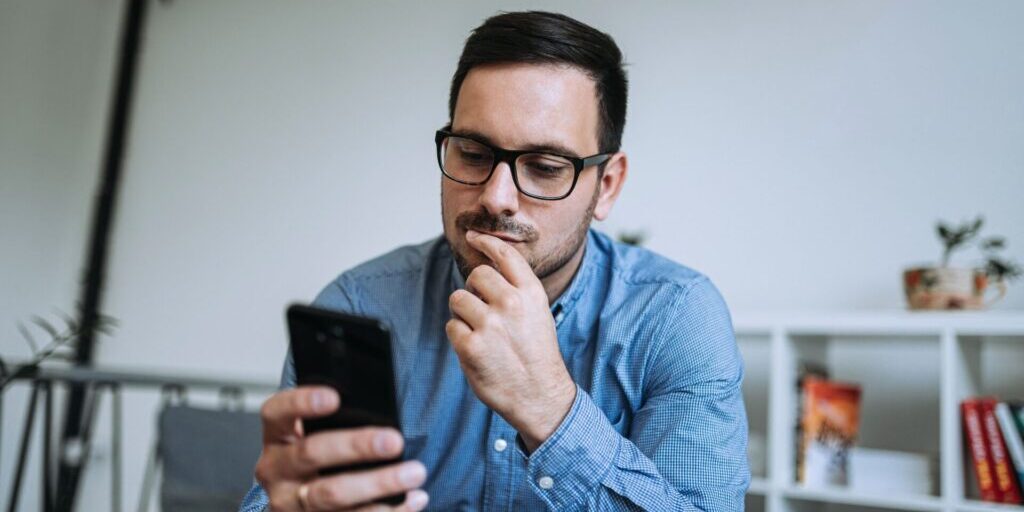 Young thoughtful man looking at mobile phone, concerned about a smishing scam.
