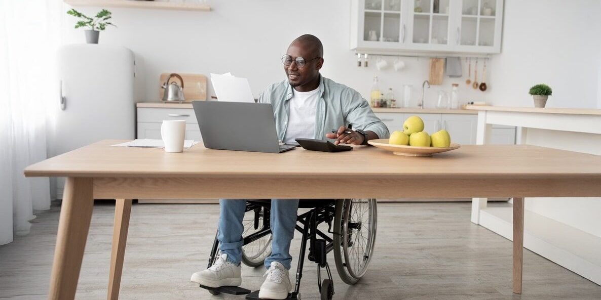A man in a wheelchair works on a laptop at a modern kitchen table with a coffee, papers, and a bowl of green apples. The scene is bright and focused.