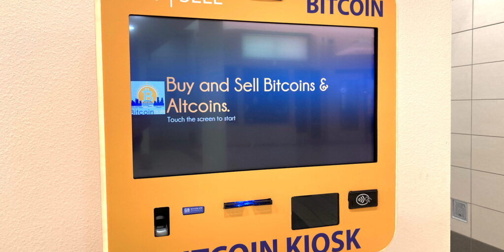 An orange Bitcoin Kiosk mounted on a light wall displays a screen prompting users to "Buy and Sell Bitcoins & Altcoins.