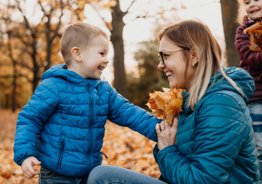 A smiling woman in a teal jacket kneels beside a boy in blue as they share autumn leaves in a park, with another child behind them.