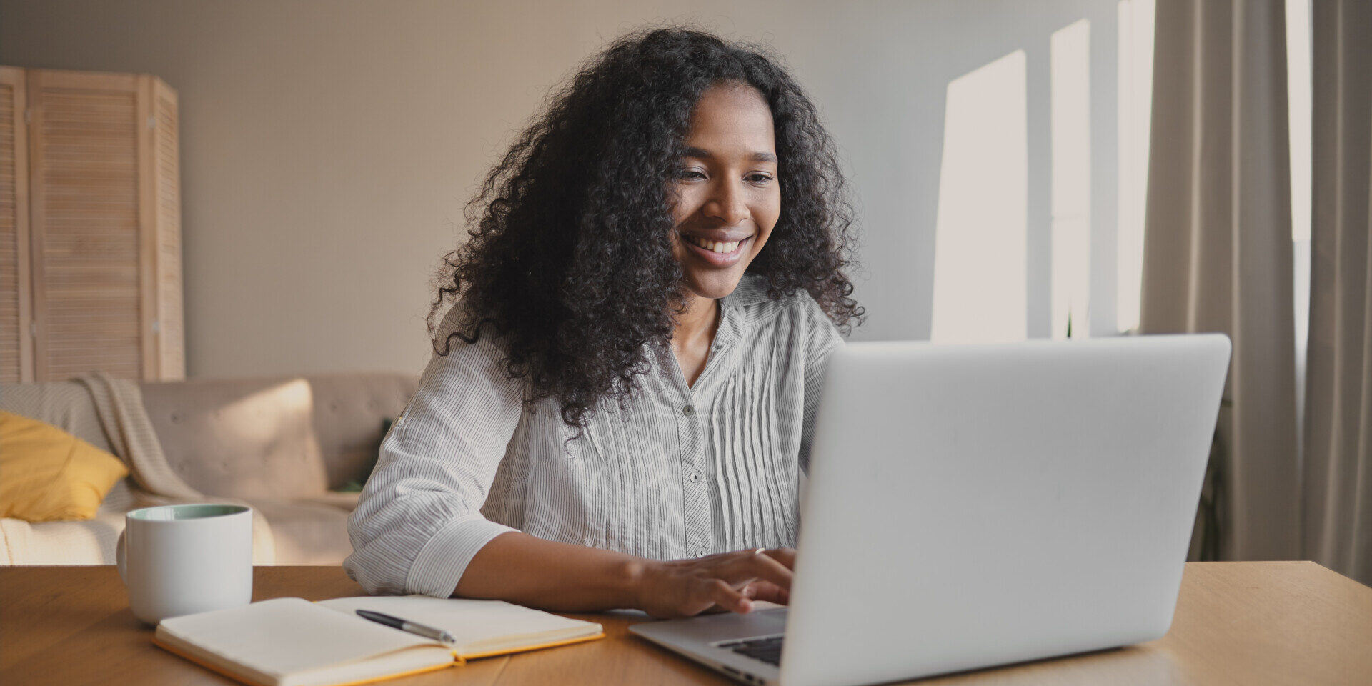 Cheerful young African American woman copywriter sitting in front of open laptop with mug and copybook on desk, checking estatements.