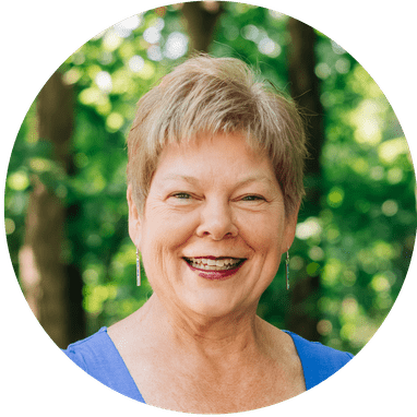 A warm, friendly portrait of Debbi Wachter, Financial Advisor at Park View Advisors, smiling. She has short gray hair and dangling earrings, wearing a blue top in a sunlit green forest.
