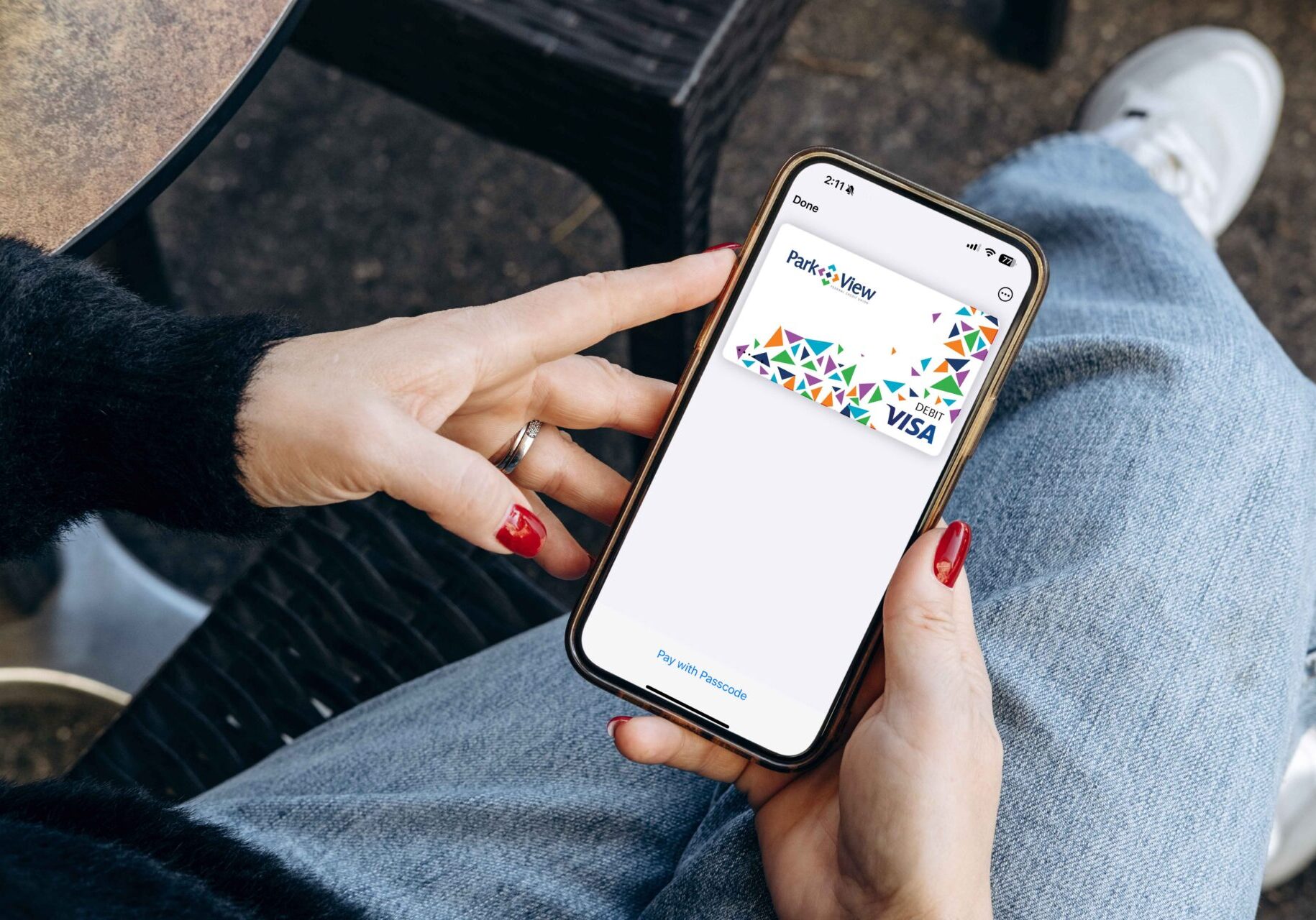 Close-up of a person holding a smartphone displaying a colorful Visa card in a banking app while seated outdoors in jeans.