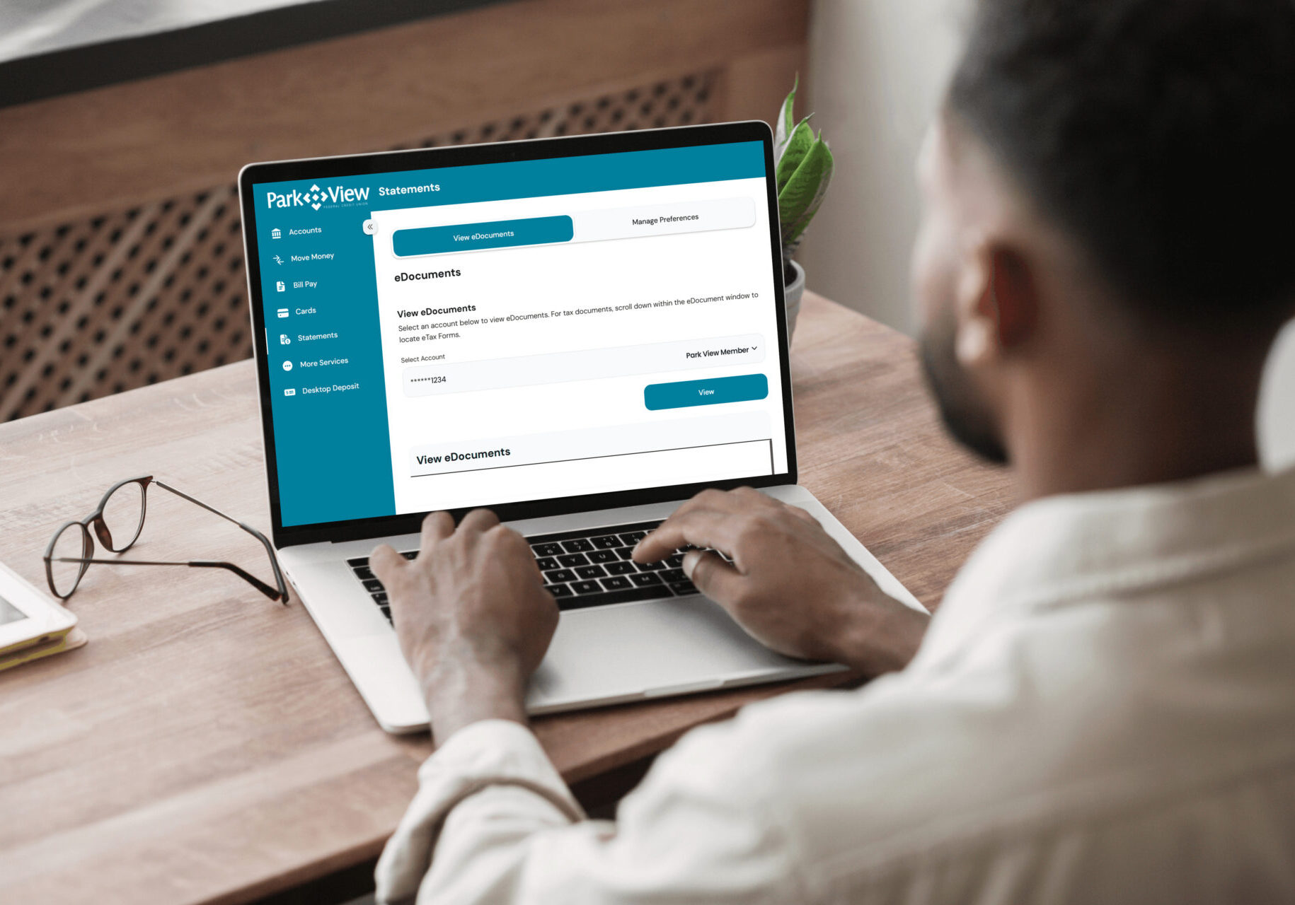 A man sits at a wooden table using a laptop displaying a financial management webpage with tabs like "eDocuments." Glasses lie beside the laptop.