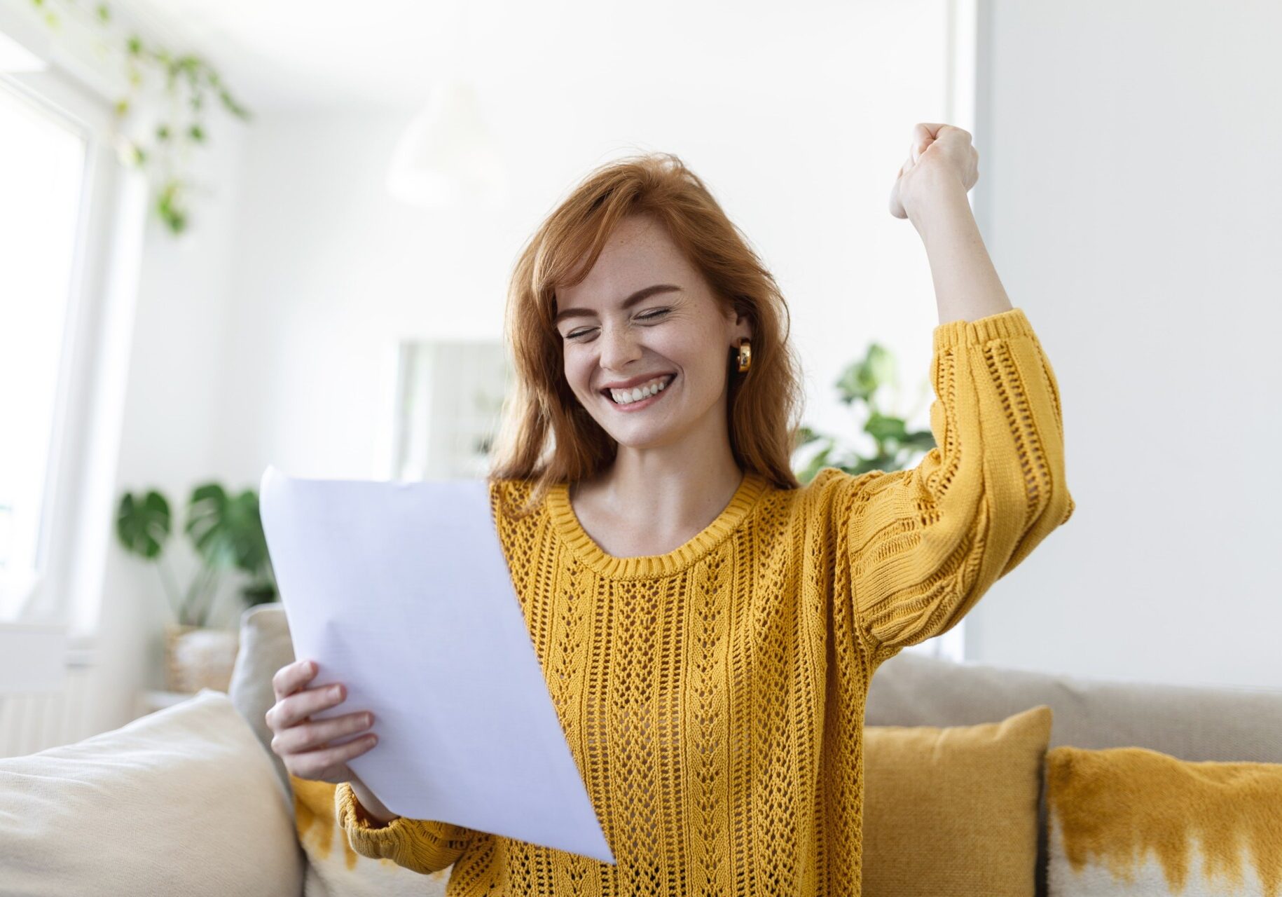 Excited, smiling young woman holds a paper with her right hand while making an excited fist motion with her left hand while sitting on a couch.