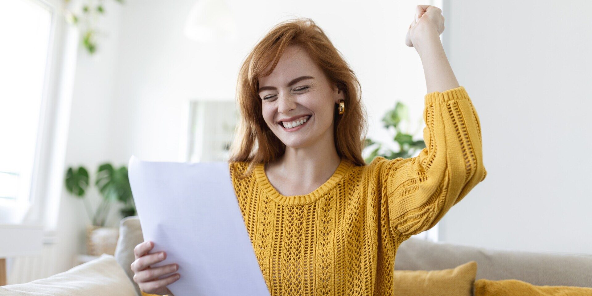 Excited, smiling young woman holds a paper with her right hand while making an excited fist motion with her left hand while sitting on a couch.