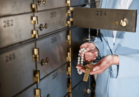 A person in a suit holds a key and a pearl necklace, standing next to a safe deposit box.
