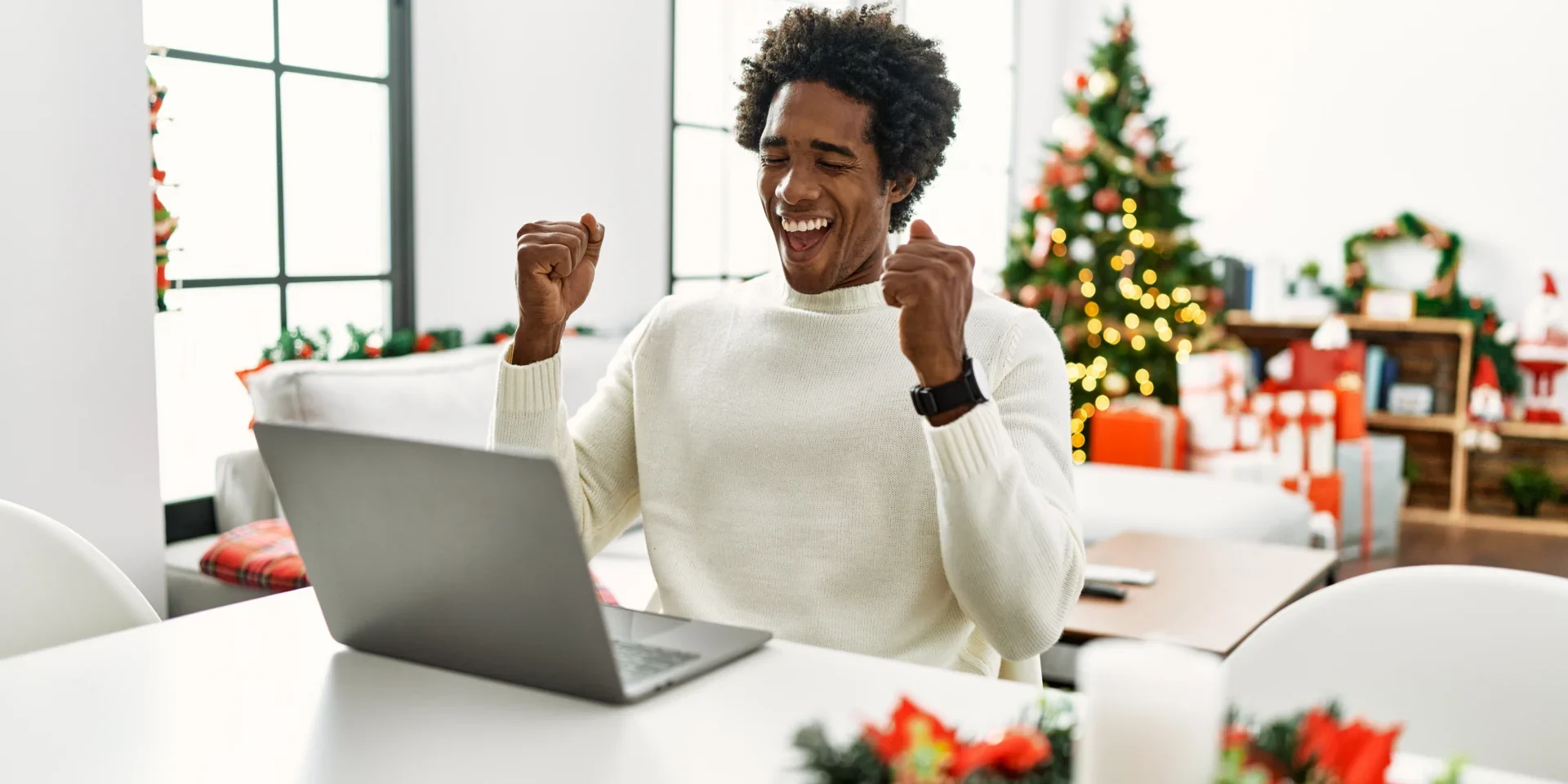 A joyful man in a white sweater raises his fists beside a laptop, celebrating in a bright office decorated with a Christmas tree.