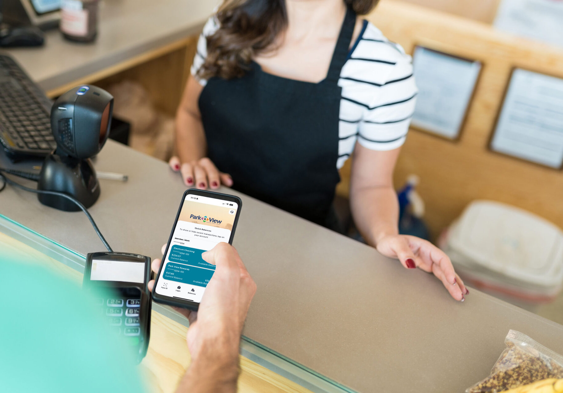 A man's hand holding a phone at a grocery store check out, using Park View's quick balance feature to check his balance.
