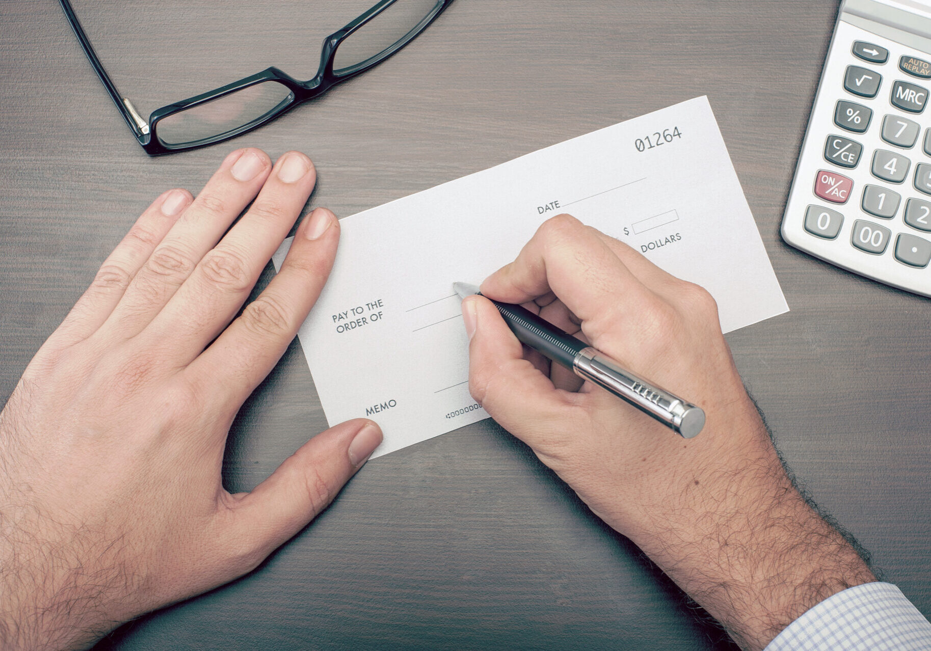 Top-down view of hands filling out a financial check with a pen on a desk, beside eyeglasses and a calculator; conveys focused money management.