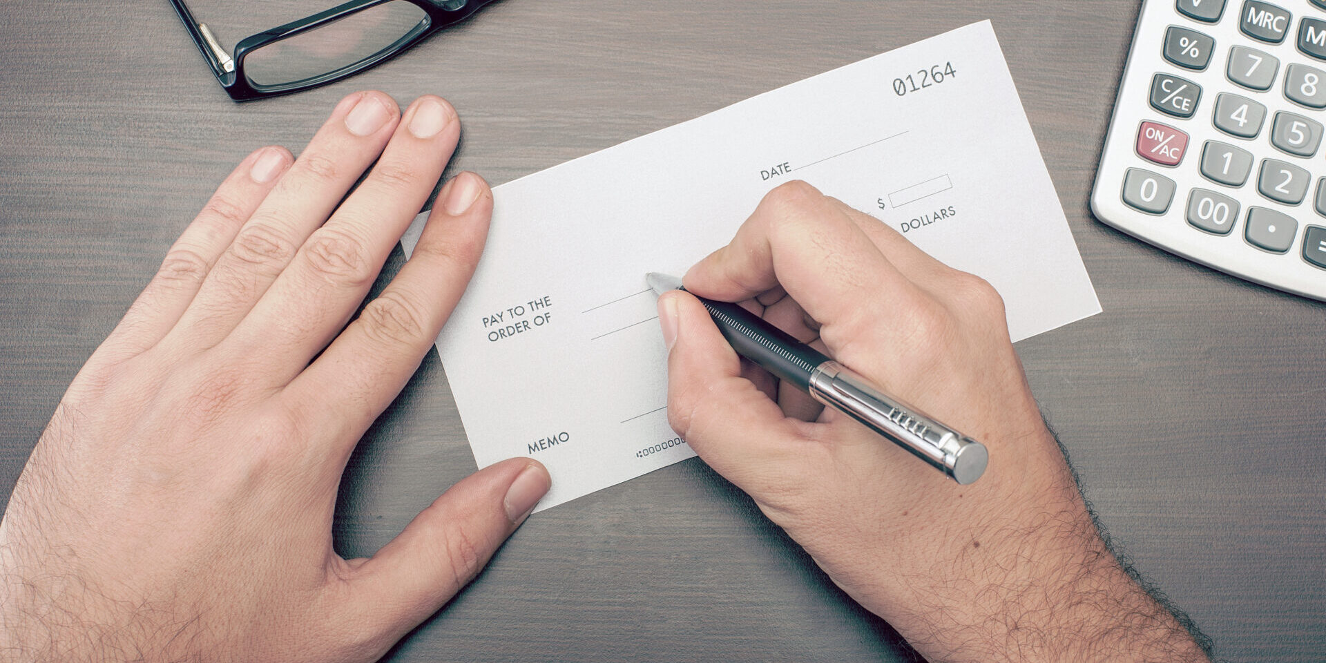 Top-down view of hands filling out a financial check with a pen on a desk, beside eyeglasses and a calculator; conveys focused money management.